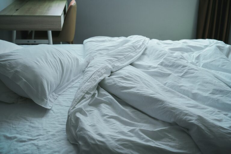 Bettwäsche wechseln – ungemachtes Bett mit weißen Laken und Nachttisch