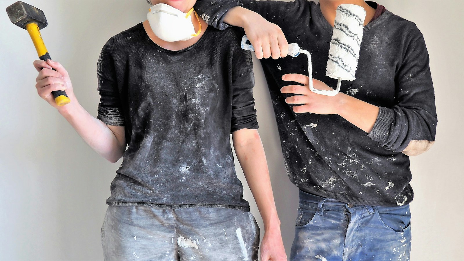 Unterschied Sanierung Renovierung: Paar mit Vorschlaghammer und Farbrolle vor einer Wand
